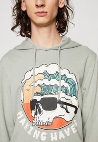 Felpa verde chiaro con cappuccio, caratterizzata da una stampa grafica di un teschio con occhiali da sole e onde, incorniciato da un cerchio arancione e dal testo "MAKING WAVES."