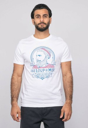 Homme aux cheveux foncés et barbe portant un T-shirt blanc avec un graphisme nautique bleu et rouge représentant un marin barbu et un texte en français.