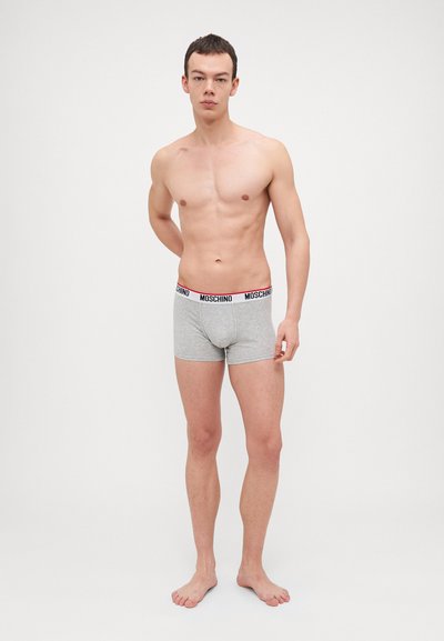Grå bomulds boxer shorts med elastisk talje, der har rød og sort "Moschino" branding. Modellen står barfodet med hænderne bag ryggen.