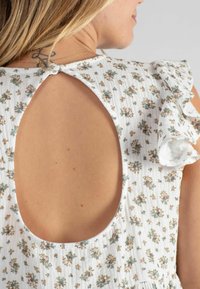 Weißes Blumenkleid mit Rüschärmeln, Schlitz im Rücken und kleinen grünen und rosa Blumenmustern auf einem weichen Textil mit Struktur.