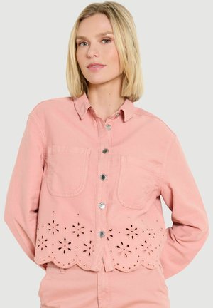 Blondínka v svetloružovej bunde so striebornými patentkami, prednými vreckami a kvetinovými výrezmi pozdĺž vlnovitého lemu.