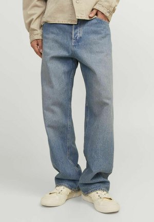 Personne portant un jean large bleu clair, une chemise beige et des baskets blanc cassé, debout avec une main dans la poche sur fond uni.