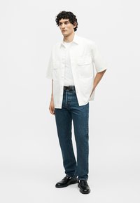 Mężczyzna o kręconych czarnych włosach, ubrany w białą, krótką koszulę zapinaną na guziki na białym T-shircie, niebieskie dżinsy, czarny pasek i czarne buty, stoi z rękami za plecami.