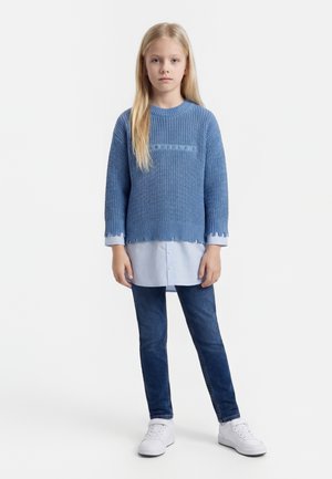 Joven chica con pelo largo y rubio que lleva un suéter de punto azul sobre una camisa azul clara, vaqueros oscuros y zapatillas blancas, de pie frente a un fondo blanco.