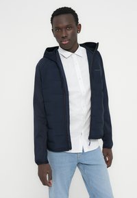 Νεαρός άνδρας με μπλε σκούρο μπουφάν με κουκούλα πάνω από λευκό πουκάμισο με σχέδια και ανοιχτό μπλε τζιν, μπροστά σε απλό φόντο.