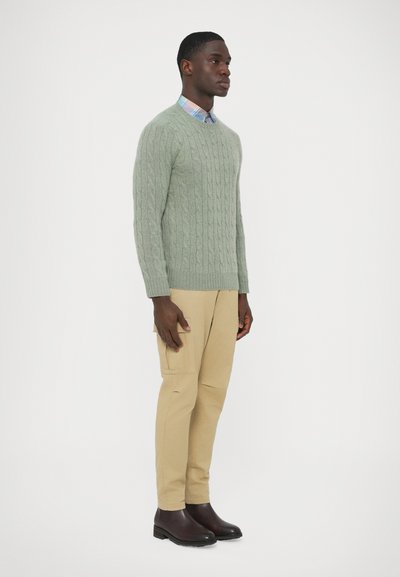 Suéter verde claro de malha trançada, calças bege e botas castanho-escuro. Camisa xadrez visível na gola, usada por baixo. Vista lateral.