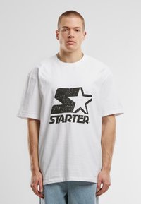 White oversized t-shirt made of cotton, featuring a black distressed "STARTER" logo with a star. Short sleeves and crew neck design.