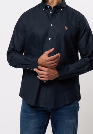 Mann trägt ein marineblaues Hemd mit Knöpfen und einem kleinen gestickten Logo auf der Brust, passt die Manschette an, kombiniert mit blauen Jeans.