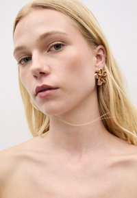 Ohrstecker in Form einer goldenen Blume mit rotem Edelstein in der Mitte. Verfügt über strukturierte Blütenblätter und eine polierte Oberfläche, vor einem neutralen Hintergrund.