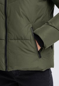 Olivgrüne Steppjacke mit durchgehendem Reißverschluss vorne, seitlichen Taschen mit Reißverschlüssen und schwarzen, gerippten Bündchen. Strukturierter Stoff mit abgesteppten Einsätzen.