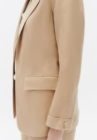Beige op maat gemaakte blazer met een gestructureerde afwerking, enkele knoopmanchet en een opgestikte zak, getoond vanuit de zijaanzicht.