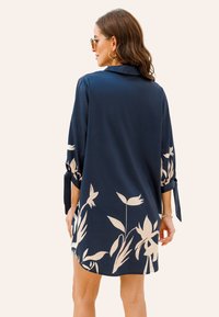 Vestido camisa azul marino con mangas de tres cuartos, que presenta un diseño floral en beige claro en los laterales y detalles atados en las mangas.