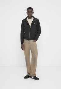 Άνδρας που στέκεται μπροστά σε λευκό τοίχο, φοράει μαύρο σακάκι με φερμουάρ, μπεζ μπλούζα, ξεθωριασμένο μπεζ τζιν και μαύρα αθλητικά με λευκές ρίγες.