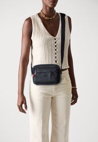 Svart crossover-väska med texturerat material, framficka med dragkedja och justerbar rem; bärs över en stickad vit topp och beige vida byxor.