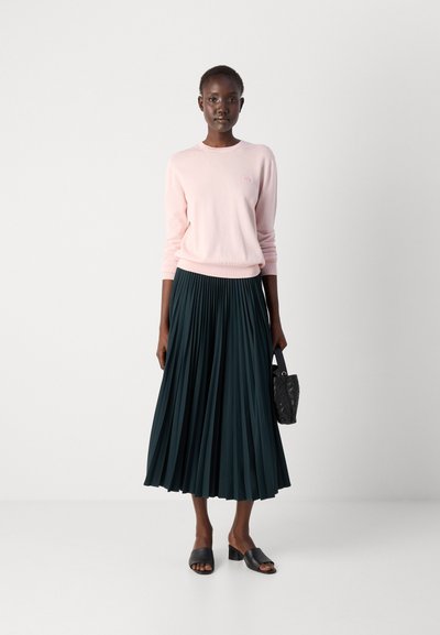 Un pull rose clair, une jupe plissée vert foncé, des mules noires à talons et un petit sac noir. Textures douces et contrastes de couleurs distincts.