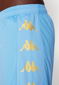 Pantalones cortos deportivos azul claro con logotipos de siluetas doradas verticales de dos figuras sentadas espalda con espalda en el panel lateral.