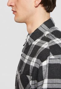 Sort-hvid ternet flannel skjorte med knapkrave, der har en brystlomme og et blødt, struktureret stof. Sideprofil udsigt.