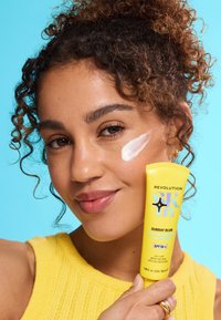 Gul tube med solkrem med teksten "Revolution Skin", som viser en jevn hvit krem på huden. Bakgrunnen er klar blå. Modellen har på seg en gul topp.