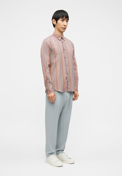 Άνδρας που στέκεται, φορώντας πουκάμισο με πολύχρωμες κατακόρυφες ρίγες και κουμπιά, ανοιχτόχρωμο γκρι χαλαρό παντελόνι και λευκά αθλητικά παπούτσια, σε απλό λευκό φόντο.