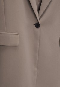 Blazer beige avec un tissu lisse, un seul bouton noir et deux poches à l'avant. Comprend un revers classique et une coupe cintrée.