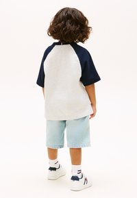 Παιδί με καστανά σγουρά μαλλιά στέκεται γυρισμένο προς τα πίσω, φορώντας γκρι και ναυτικό μπλουζάκι, ανοιχτόχρωμο τζιν σορτς, λευκές κάλτσες και λευκά αθλητικά παπούτσια.