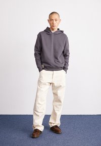 Hanorac gri cu buzunar tip cangur și glugă cu șnur, asortat cu pantaloni largi crem. Pantofii maronii au un design franjurat.