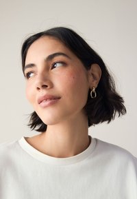 Gold hoop earrings worn by a person with short, dark hair. White t-shirt. Subtle freckles and natural makeup on face. Neutral background.