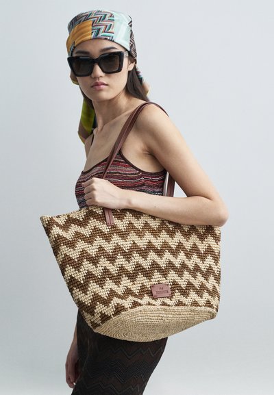 Tkaná torba z jute s hnědým a krémovým cikcak vzorem, s hnědými koženými uchy a malou koženou nášivkou s logem.