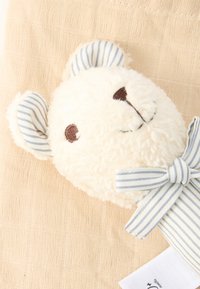 Κεφάλι λευκής πολυτελούς αρκούδας με καφέ κεντημένα μάτια και μύτη, αυτιά και φιόγκο με ριγέ μπλε και λευκό, σε μπεζ υφή υφάσματος.