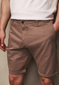Pantalones cortos de algodón marrón con corte recto, con cierre de botón, puños doblados y bolsillos laterales. Textura suave y diseño minimalista.