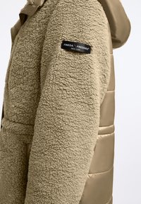 Beżowy, teksturowany płaszcz z owczej wełny o błyszczącym, satynowym wykończeniu. Posiada kaptur, boczne kieszenie oraz czarną łatkę z logo na rękawie.