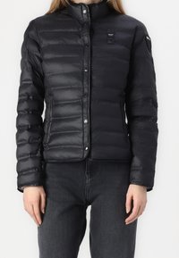 Sort pufferjakke, letvægt, med vandret quiltet design, trykknapper og høj krave. Båret over mørke denimbukser.