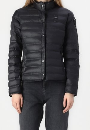 Zwarte pufferjack, lichtgewicht, met horizontaal gestikte motief, drukknoopjes en hoge kraag. G gedragen over donkere spijkerbroek.