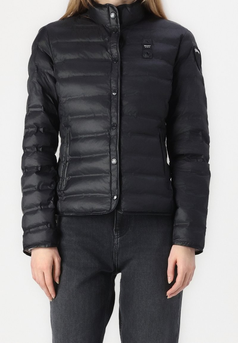 Sort pufferjakke, letvægt, med vandret quiltet design, trykknapper og høj krave. Båret over mørke denimbukser.