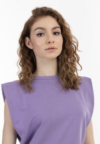 Lilla ærmeløs top med strukturerede skulderpuder og en blød tekstur. Modellen har bølget hår og minimal makeup. Baggrunden er hvid.