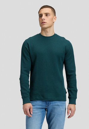 Mann trägt einen dunkelgrünen, langärmeligen, strukturierten Pullover und hellblaue Jeans und steht vor einem schlichten Hintergrund.