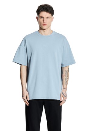 Młody mężczyzna z krótkimi ciemnymi włosami, ubrany w jasnoniebieski oversize T-shirt i czarne spodnie, pokazujący tatuaże na obu przedramionach i dłoniach.