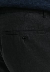 Schwarze Leinenhosen mit einer Gesäßtasche, die einen einzelnen schwarzen Knopf aufweist. Der Stoff hat eine dezente Textur, die zu einem dunkelblauen Oberteil passt.