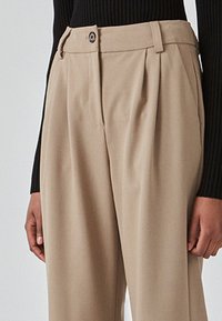 Beige bukser med pleats, der har en knaplukning og bæltestropper. Stoffet har en glat tekstur og vises sammen med en sort ribbet top.