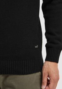 Black knitted sweater with ribbed hem and cuffs. Small brand tag on the lower side. Smooth texture and tailored fit.