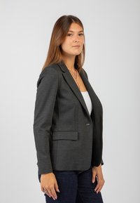 Blazer grigio scuro con un design aderente, chiusura a bottone singolo e due tasche frontali. Abbinato a un top bianco e jeans scuri.