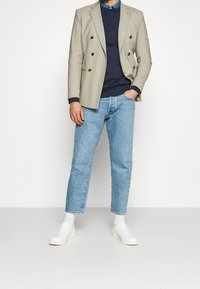 Beige Doppelreiher-Blazer über einem marineblauen Sweater, kombiniert mit hellblauen Jeans und weißen Sneakers, was einen lässigen, aber dennoch eleganten Look zeigt.