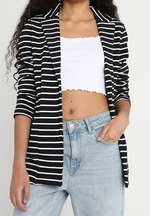 Kvinde iført sort- og hvidstribet blazer, hvid cropped top og lyseblå højttaljede jeans med den ene hånd i lommen.