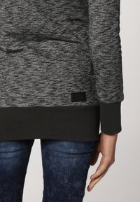 Szara melanżowa bluza z czarnym wykończeniem i mankietami, teksturowany materiał oraz mała czarna metka w lewym dolnym rogu.