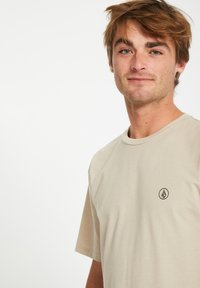 Beige t-shirt met korte mouwen van zachte stof, met een klein logo op de linkerkant van de borst. Ronde halslijn, relaxed fit, minimalistisch ontwerp.