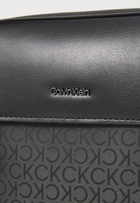 Borsa in pelle nera con logo Calvin Klein embossato; presenta una texture liscia e un motivo CK ripetuto, in contrasto con una finitura lucida.