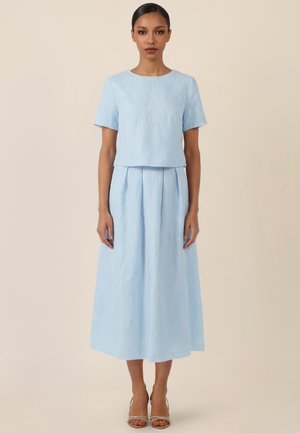 Frau mit zurückgestyltem dunklem Haar, trägt ein strukturiertes hellblaues Kurzarmoberteil und einen passenden plissierten Midirock, steht aufrecht, neutraler Hintergrund.