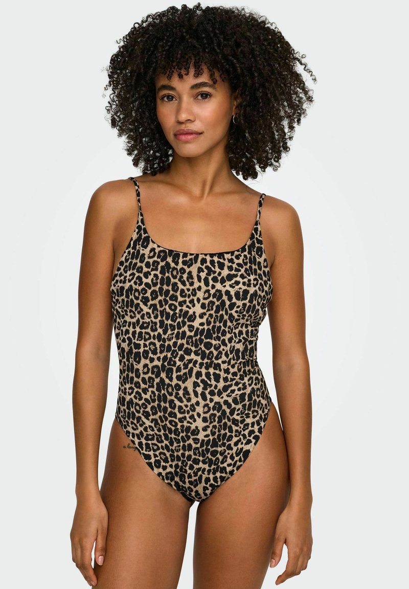 Kohaga naisega, kellel on lokkis juuksed ja kes kannab varrukateta leopardimustriga üheosalist ujumistrikood, seisab ühevärvilise heleda tausta ees.