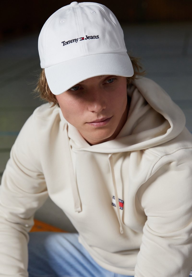 Jeune personne portant une casquette blanche Tommy Jeans et un sweat à capuche crème avec logo, regardant légèrement vers le bas dans une lumière intérieure douce.