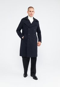 Marineblauwe dubbelgevoerde overjas met grote knopen, brede kraag en zijzakken, gecombineerd met zwarte broeken en leren schoenen.